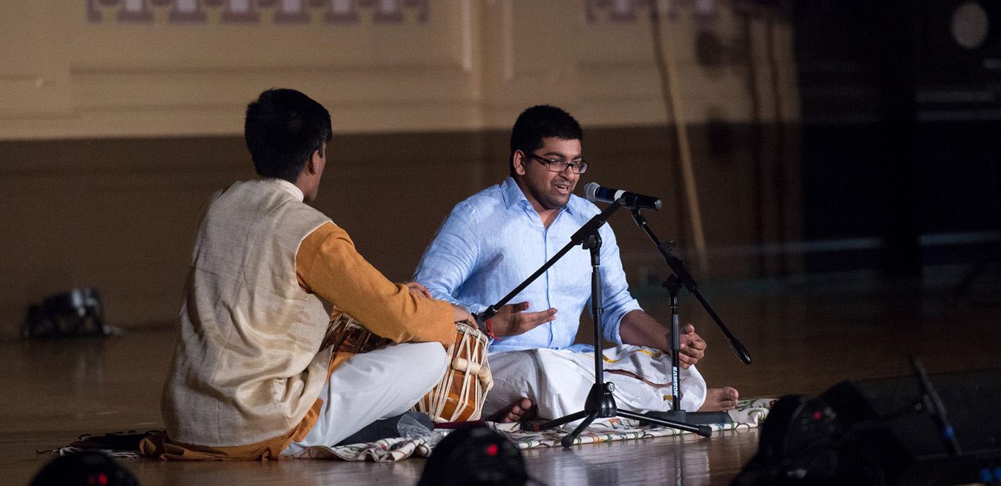 One person drums and another sings while sitting on the floor in front of microphones