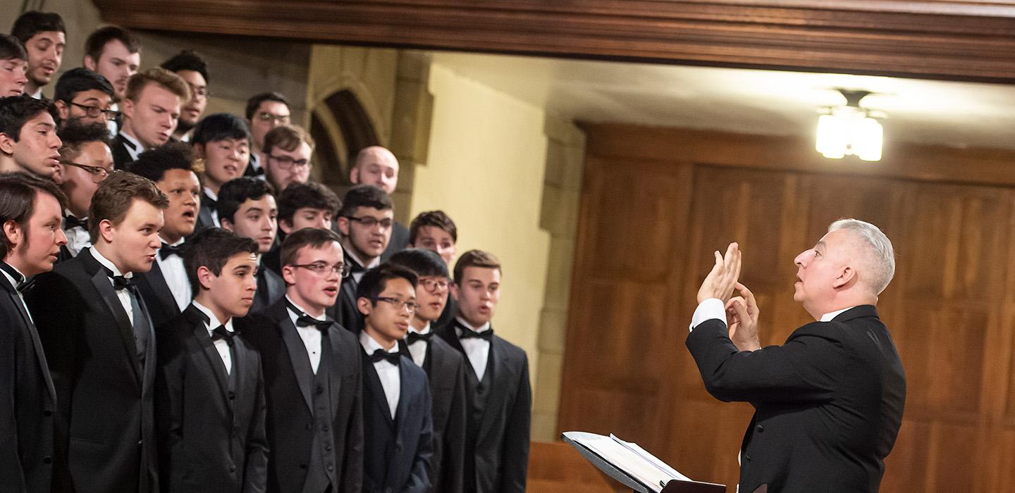 A choir of men in tuxedos are conducted