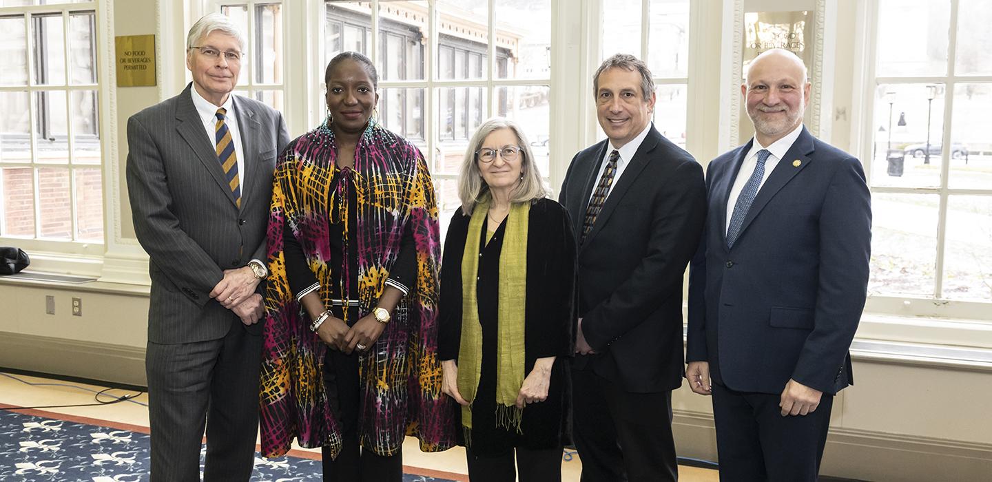 Three people in suits stand with a person in a colorful shawl and a person in a black shirt and green scarf.