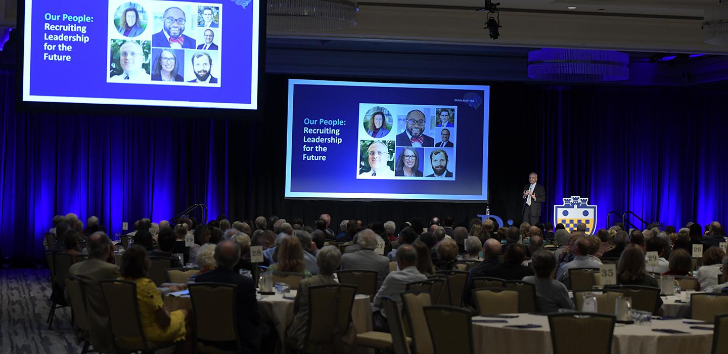 People sit at tables in a ballroom watching a presentation