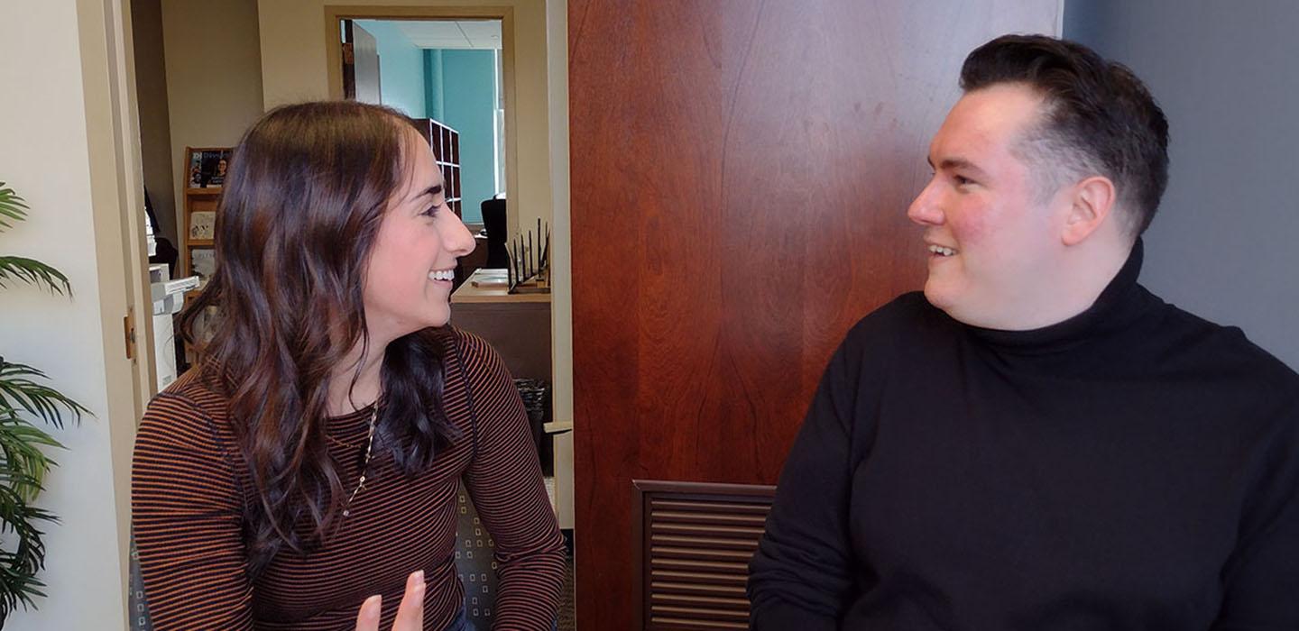 A person in a brown shirt smiles at a person in a black turtleneck