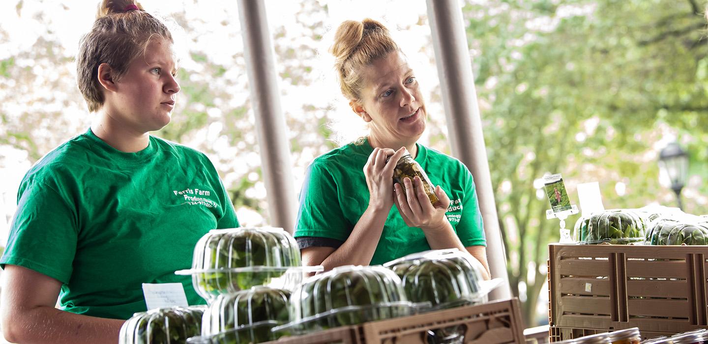 Two people in green shirts stand behind crates of greens in plastic containers