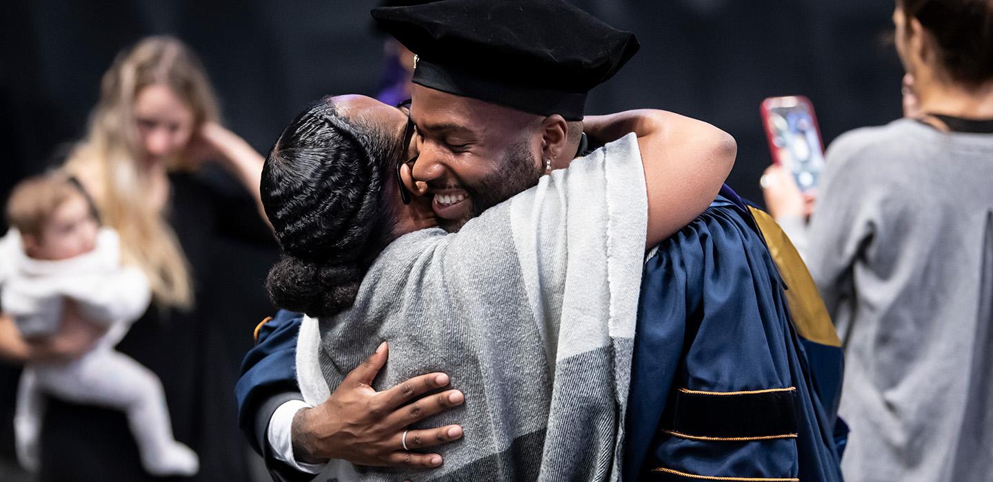 A person in blue doctoral regalia hugs someone in a grey shawl