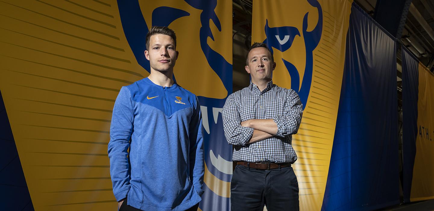 A person in a blue shirt and a person in a checkered shirt stand in front of a yellow and blue panther banner