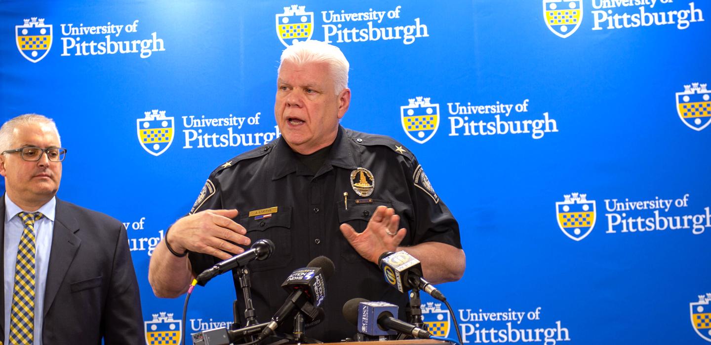 James Loftus speaks into microphones at a podium in a black Pitt Police uniform