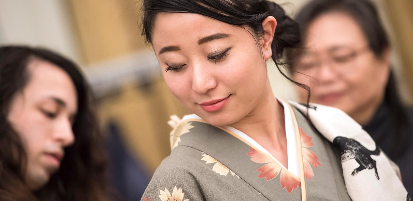 Two people stand behind a girl in a kimono. Off-camera, they are tying a ceremonial sash.