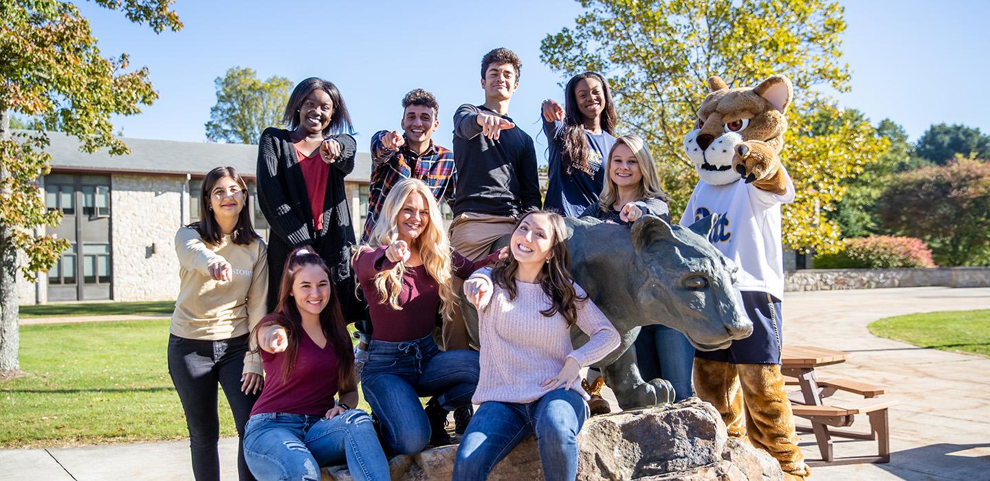 A group of students and a mascot sitting around a panther statue point at the camera