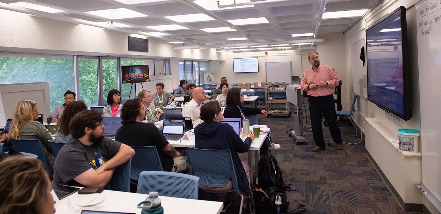 People sit with laptops in a classroom, watching a person present on a large screen