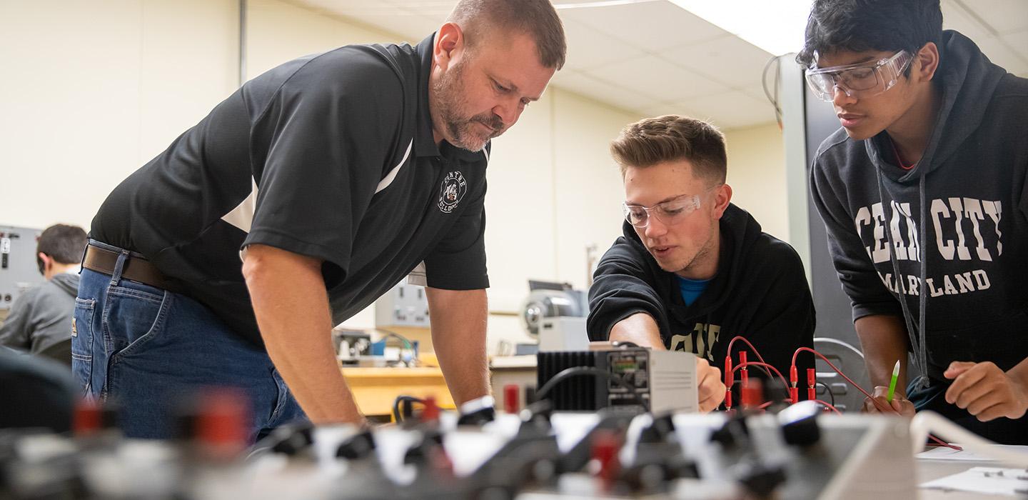 A professor talks to two students working on electrical equipment