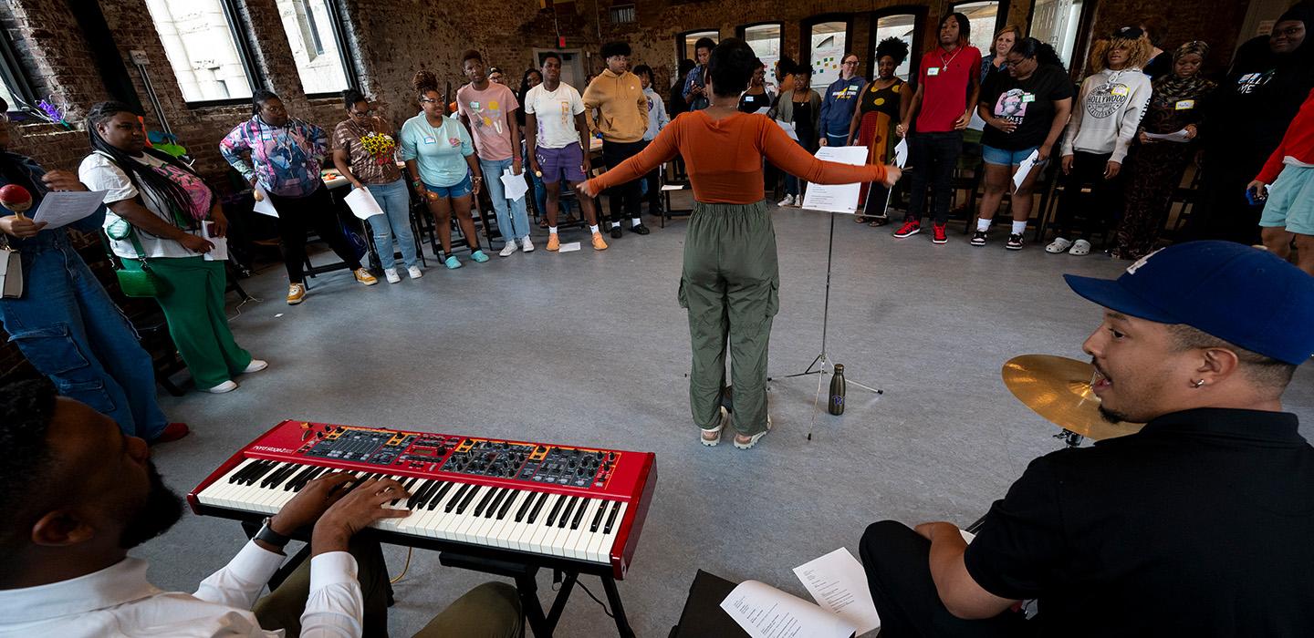 A person spreads their arms at a microphone in front of a crowd. Two musicians sit playing a keyboard and drumset behind her.