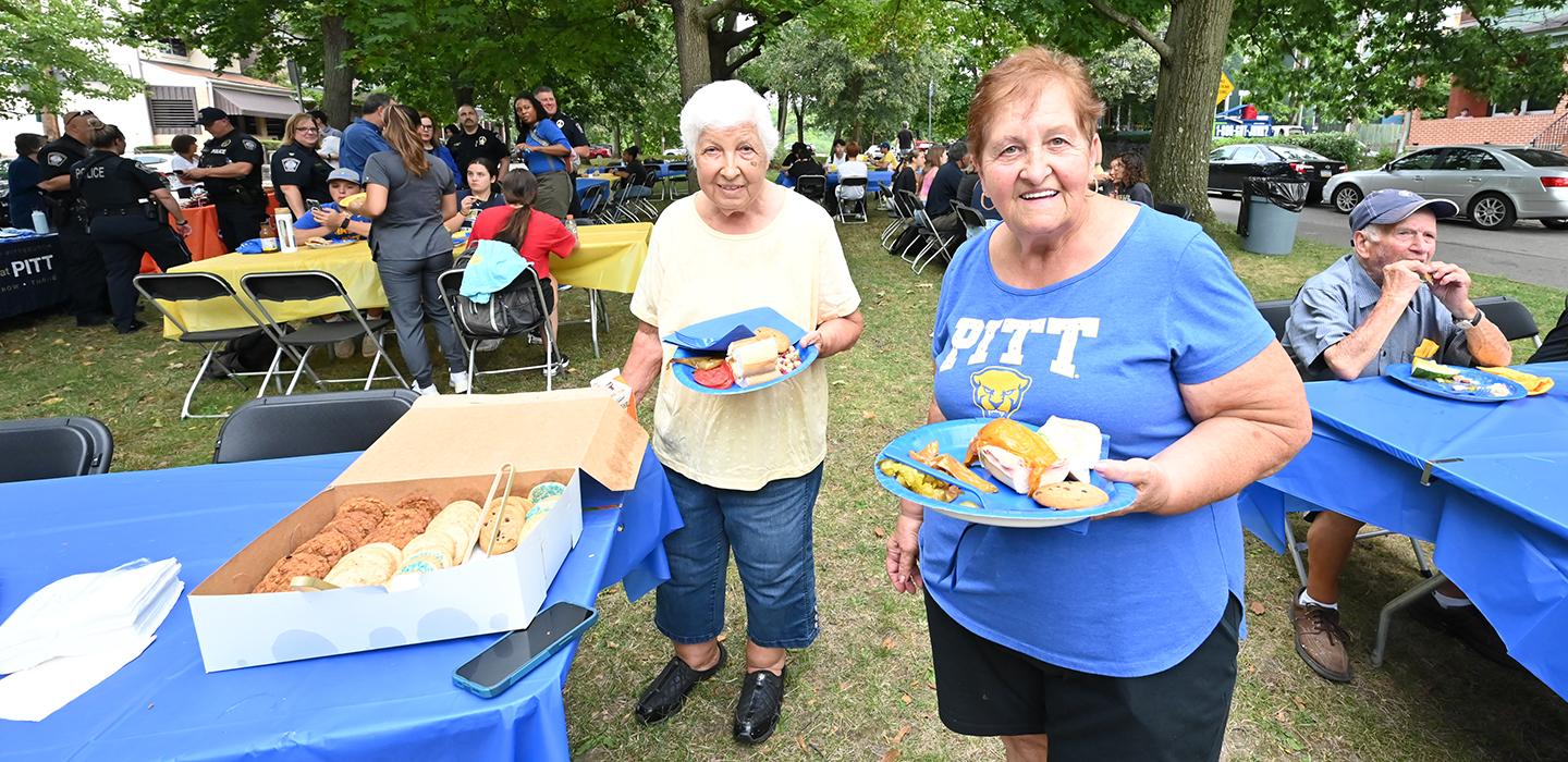 Two people fill paper plates with food. More people eat at outdoor tables in the background