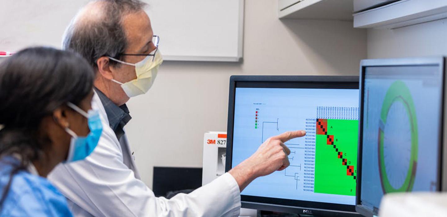A masked scientist in a white coat points at a graph on a computer screen