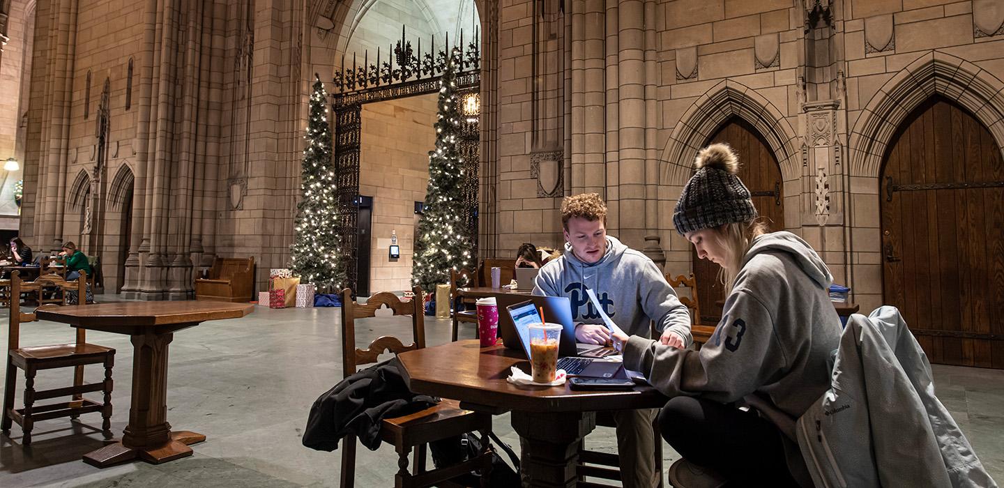 Two students study at a table in the Cathedral of Learning Commons Rooms. Two Christmas trees stand by the entrance.