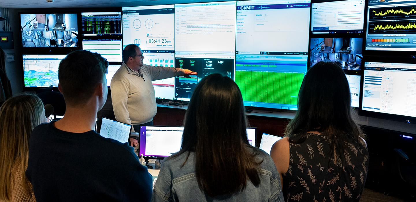 Four students watch as an instructor points at a wall of screens displaying data