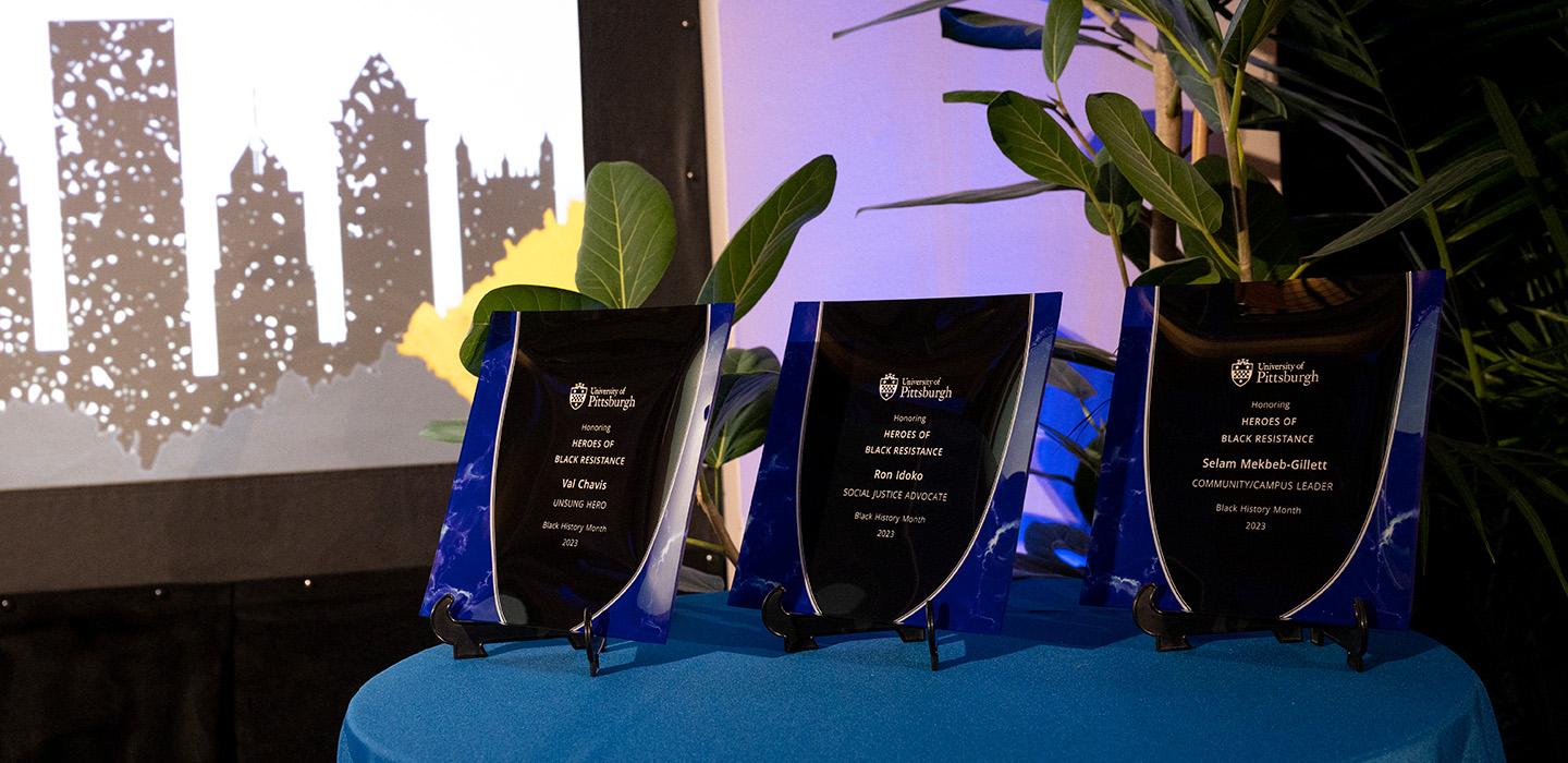 Three plaques sit on a table with a blue tablecloth in front of a projection of Pittsburgh's skyline