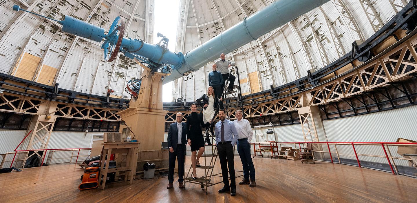 People stand on steps in a room housing a massive blue telescope