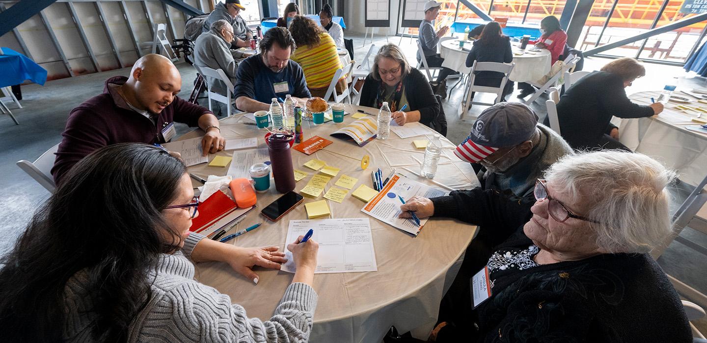 People sit at circular tables, taking notes in small groups