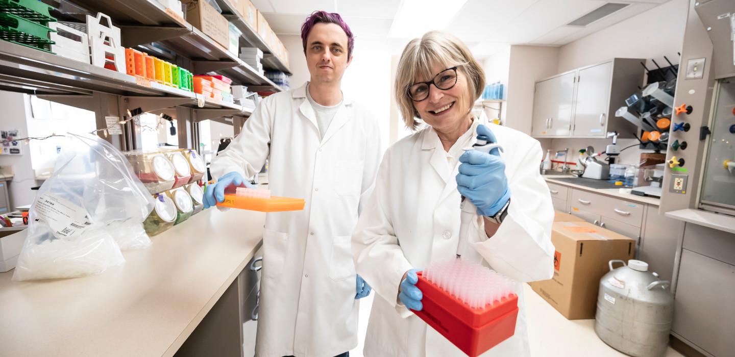 Zak Hutchinson and Anna Marsland in white lab coats, holding pipette tip boxes.