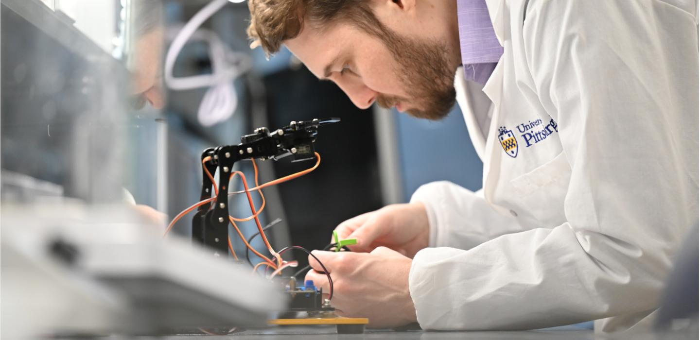 Man wearing Pitt-branded lab coat adjusts wires on electronic device