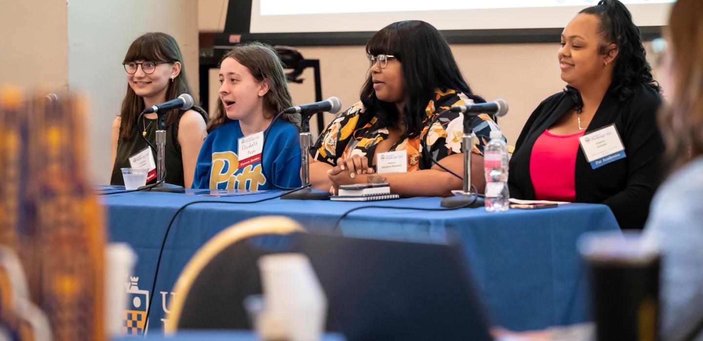 A panel of four people sit at a table with microphones