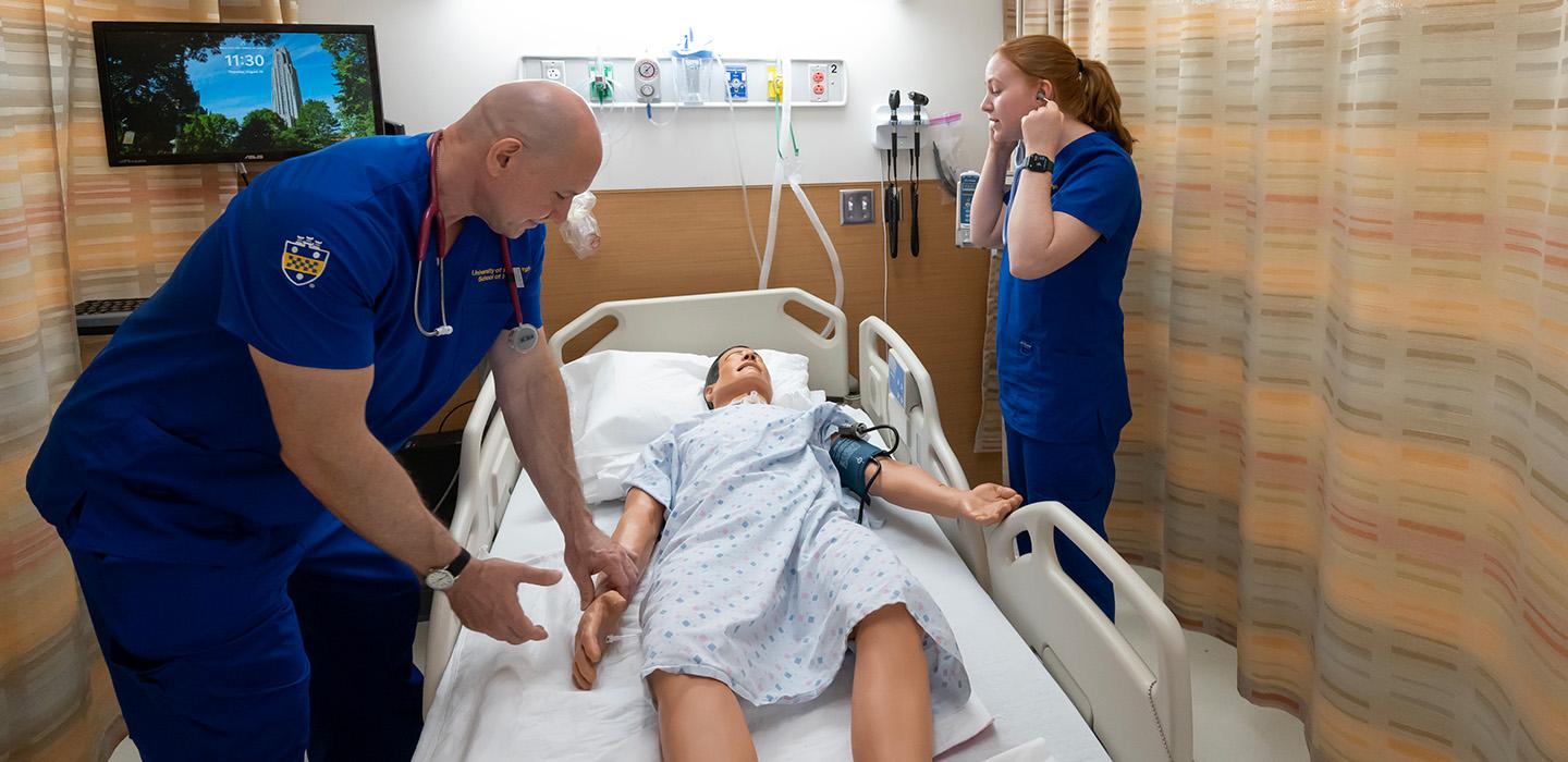 A professor demonstrates taking a pulse on a dummy in a hospital bed