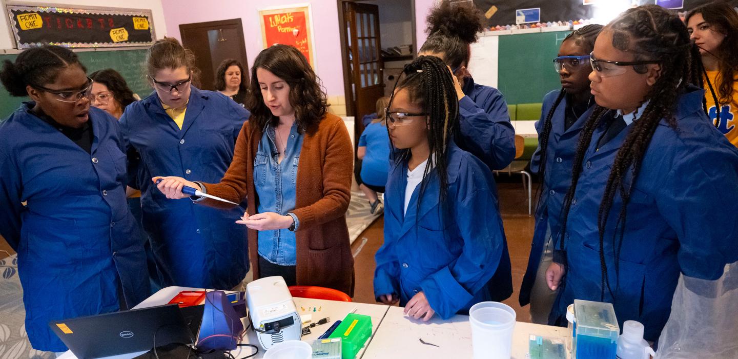 Students in blue lab coats watch as an instructor demonstrates a micropipette activity