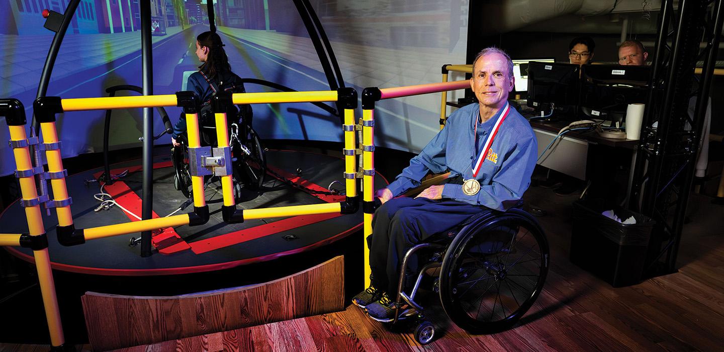 Cooper smiles next to a simulation device, which a person in a wheelchair is using. Cooper is wearing a medal.