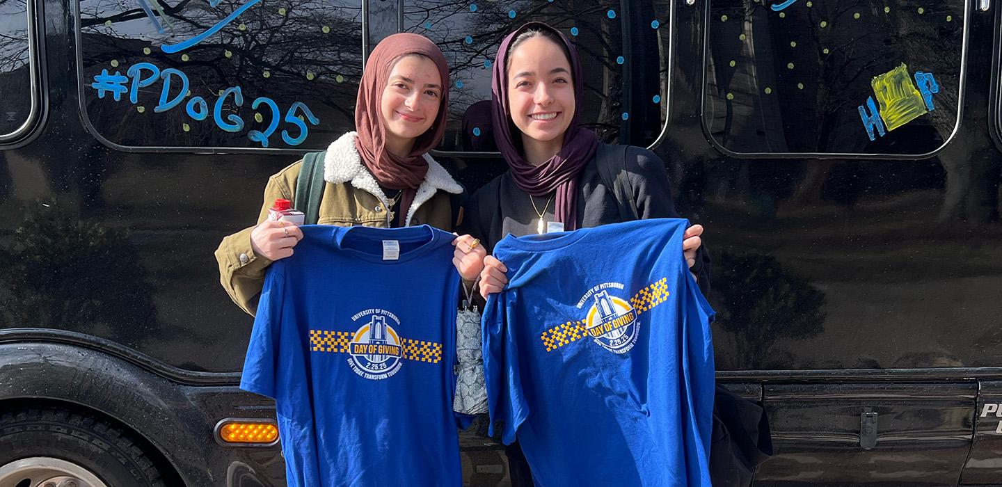 Two people hold up Pitt Day of Giving shirts in front of a black shuttle bus