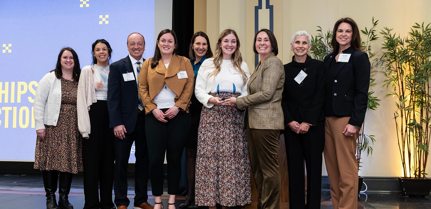 A line of awardees smiles in front of a Pitt-branded screen
