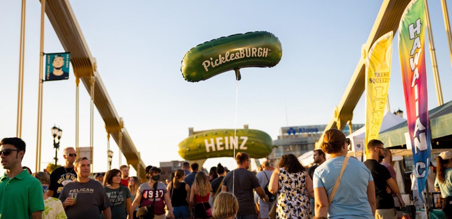 Many people on one of the Pittsburgh bridges, a child carries a pickle-shaped balloon that reads "PicklesBURGH".  In the distance there is a pickle-shaped sign reading "HEINZ".