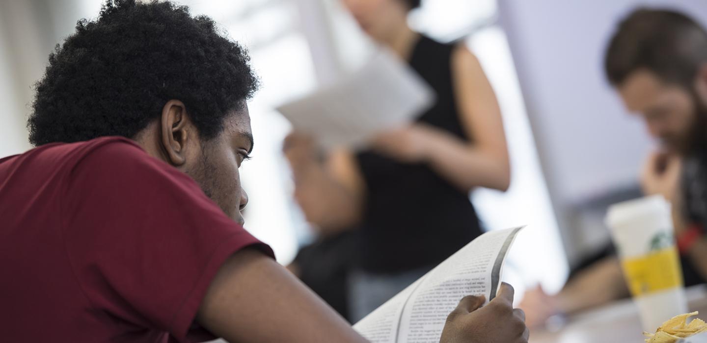 A student in a red shirt reads a book with a blurred teacher in the background.