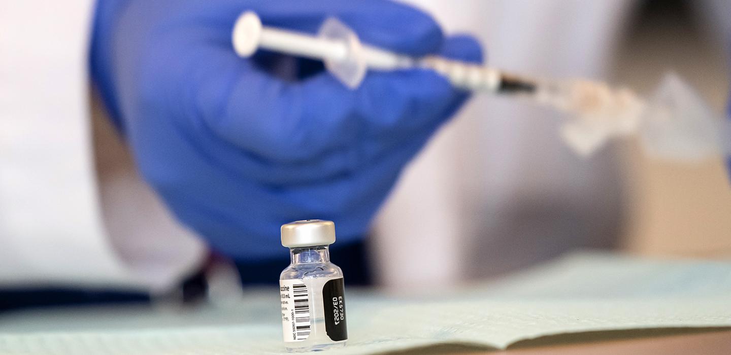 A vaccine bottle on a table with a blue gloved hand holding a syringe in the background