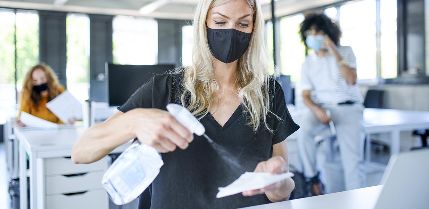 A person in a dark shirt and face mask uses a spray bottle to clean a surface