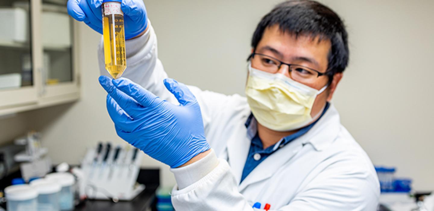 A person in a face mask, white coat and blue gloves holds up a vial