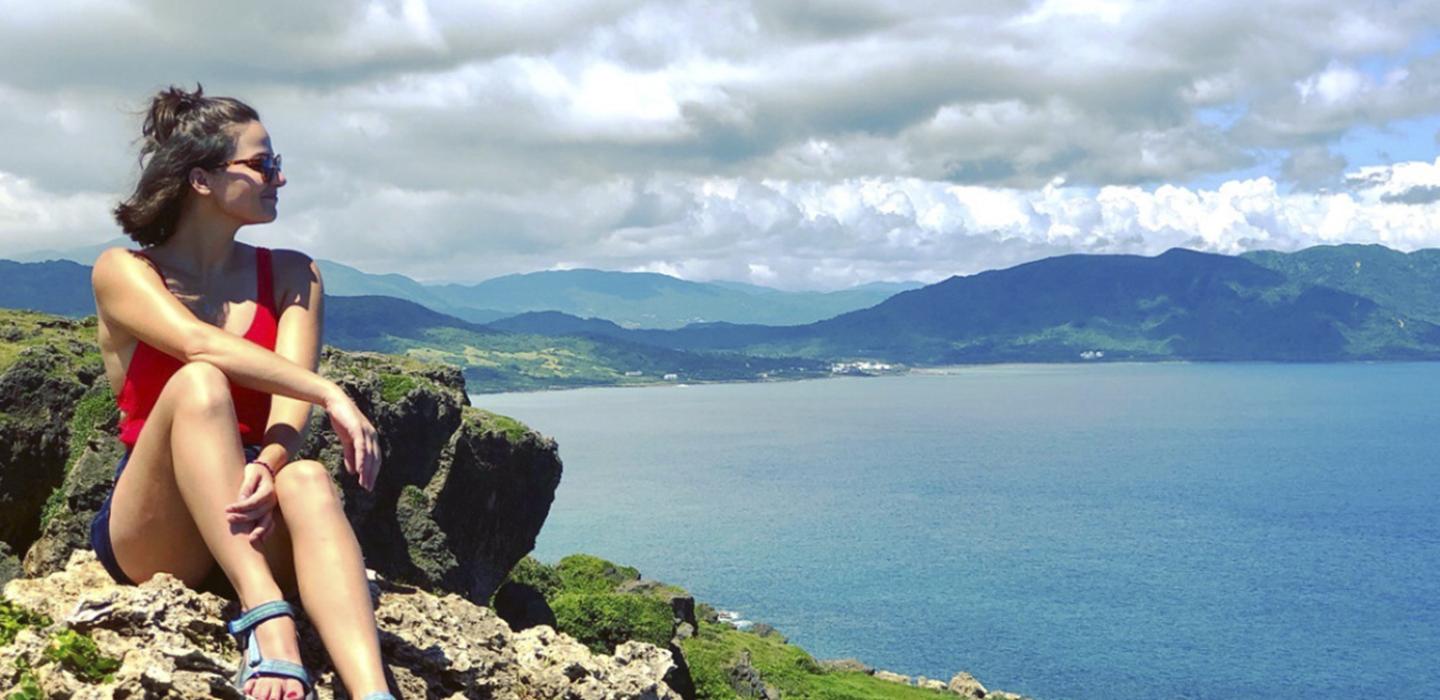 Juliette Rihl pictured seated in shorts and tank top in front of a view of mountains, water and cloudy sky