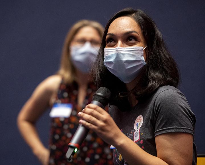 Woman speaking into microphone with mask, masked woman in background