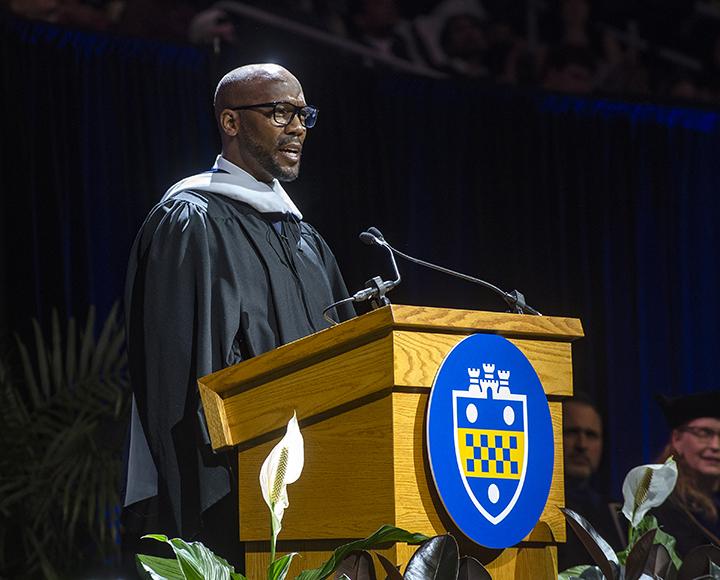 Louis Riddick in gown speaking at podium during ceremony.