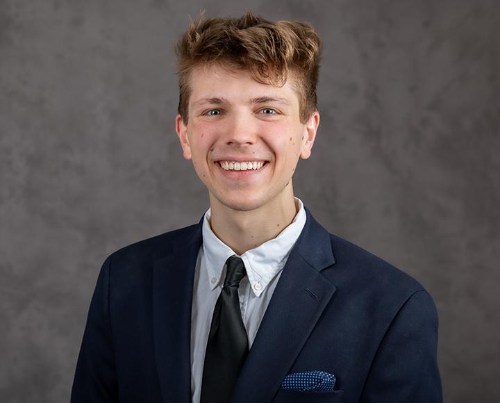 Tyler Viljaste smiling in navy blue suit in front of grey background