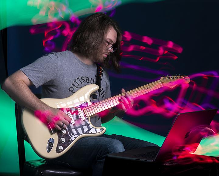 A person looks at a laptop while playing an electric guitar.