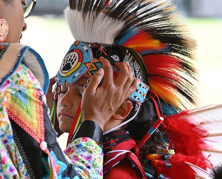 A child closes their eyes as an adult places a colorful, feathered headpiece on their head.
