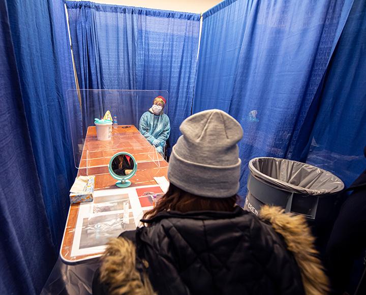 A person in a beanie sits at the far end of a table from a person in blue scrubs and a mask