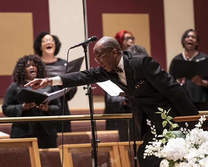 A person in a black suit conducts a choir