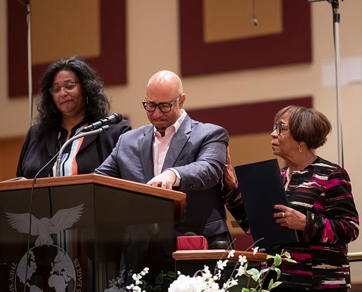 A person in a grey suit and glasses cries at a podium while a person holding a folder puts her hand on his arm.