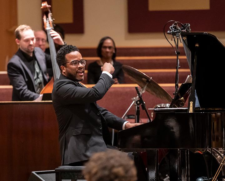 A person in a black suit plays piano in a church. An upright bassist plays behind him.