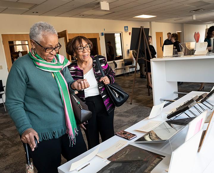 A person in a green sweater and scarf looks at materials on a table with a person in a white shirt and black jacket