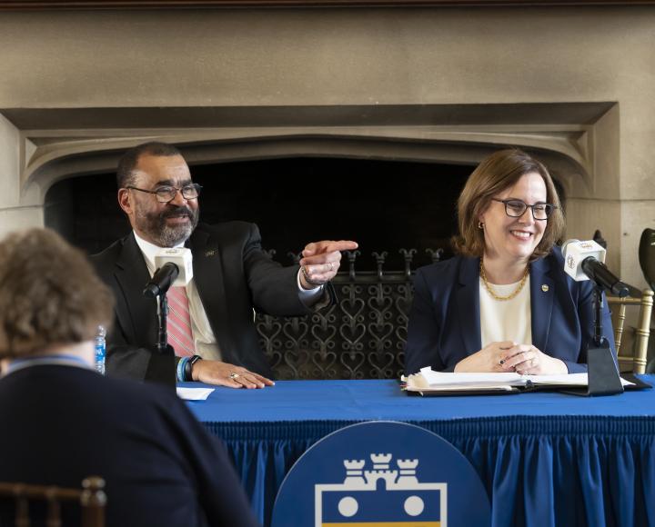 A person in a black suit and pink tie points while seating with a person in a blue suit behind a blue table