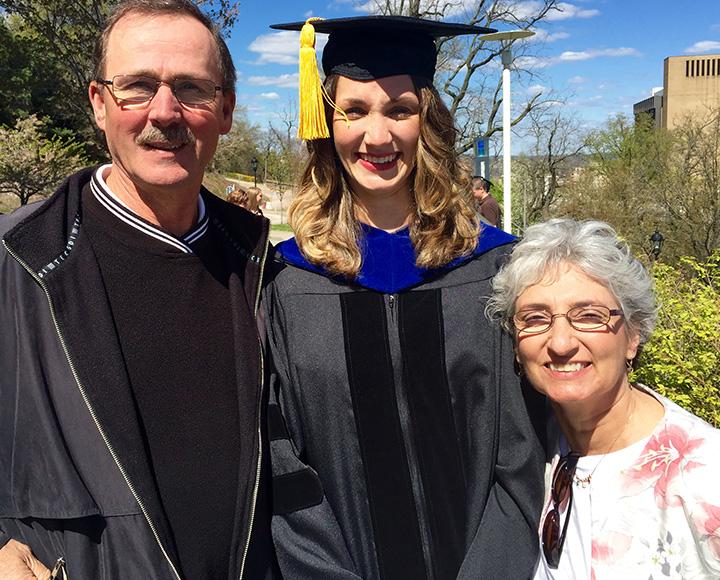 Three people smile, the person in the middle is wearing graduation regalia