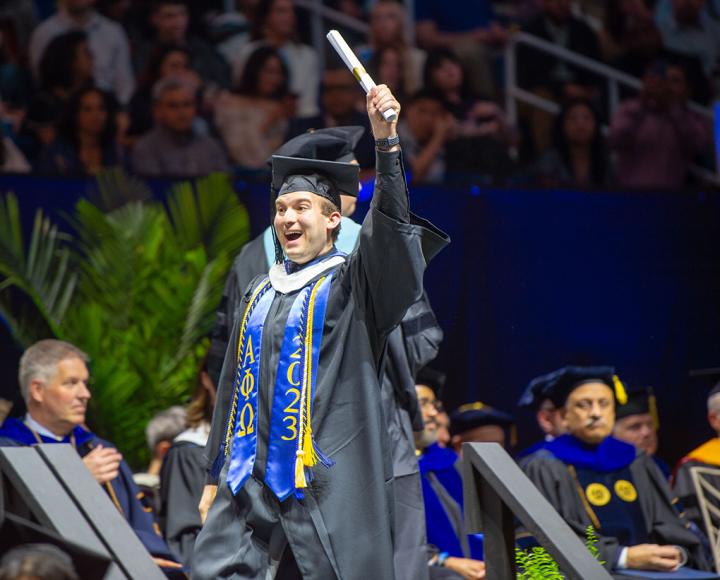 A person in black robes and a blue stole holds up a rolled up diploma