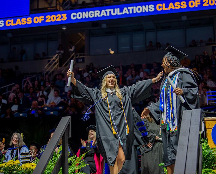 A person in graduation regalia holds up a rolled up diploma