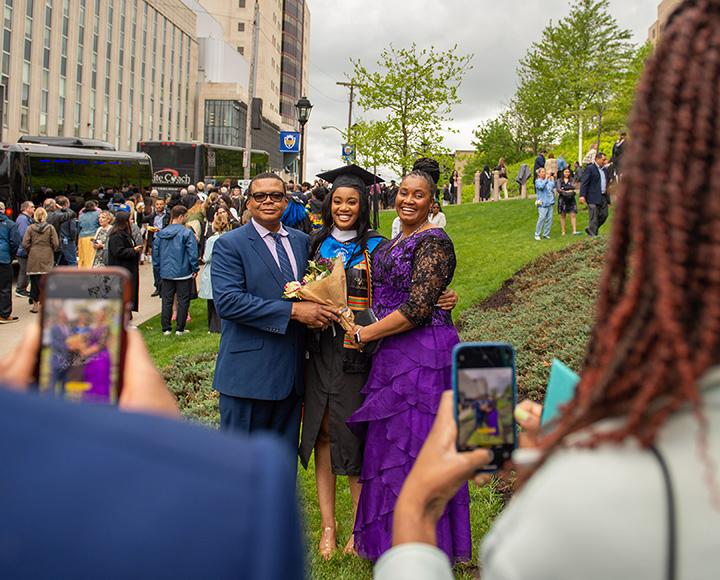 A person in a blue suit and another in a purple dress hug a person in graduation regalia